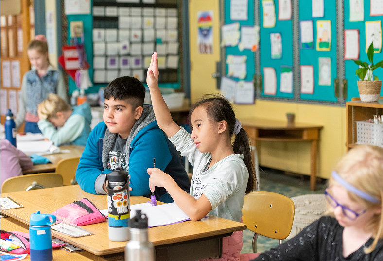 In a bright elementary classroom, a girl seated at her desk raises her hand to answer a question while the boy beside her and other classmates work in the background.