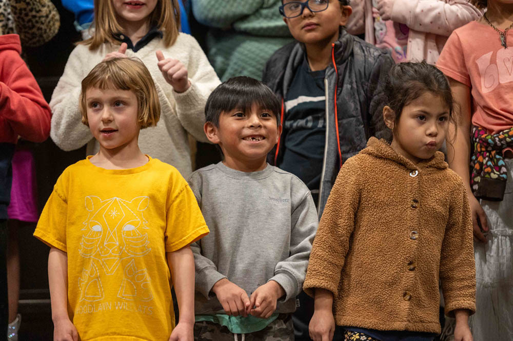 Young children stand on stage in colorful clothes, performing during a school assembly.