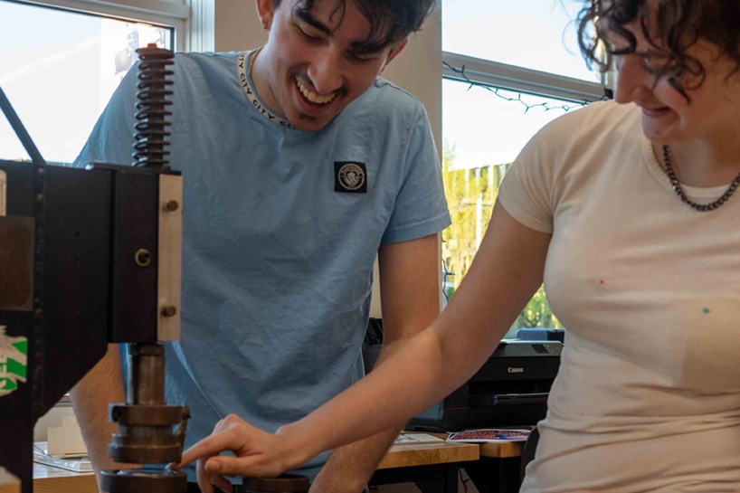Two teens smile as they operate a mechanical press in a workshop