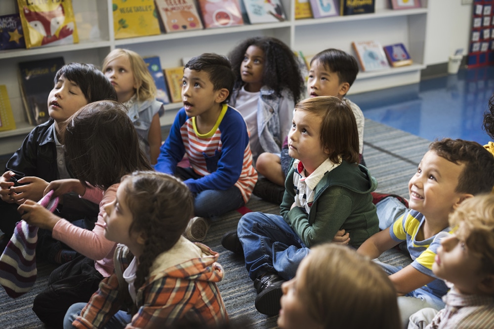 Young children sit on a classroom rug, looking up and listening attentively.