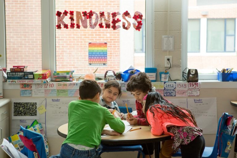 Four elementary students work together at a round table under a classroom window decorated with the word “KINDNESS.”