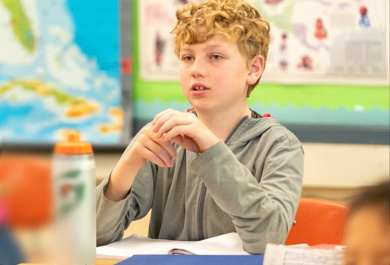 A boy sits at his desk listening during a lesson, with a world map on the bulletin board behind him.
