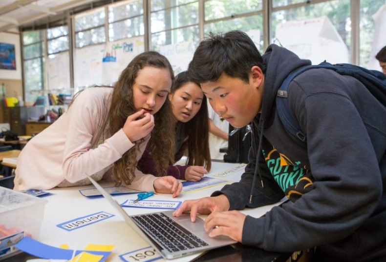 Three middle-school students gather around a laptop, collaborating on a project.
