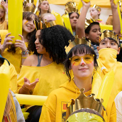 Students dressed in yellow, wearing gold paper crowns, cheer in a lively school spirit assembly.