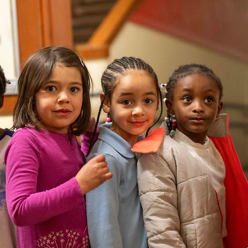 Three young girls stand in line indoors, looking toward the camera with friendly expressions.