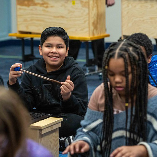 A smiling boy plays a xylophone with mallets during music class while classmates focus nearby.
