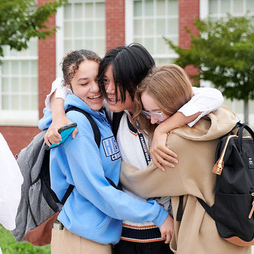 Three high-school friends share a joyful group hug outside a brick school building.