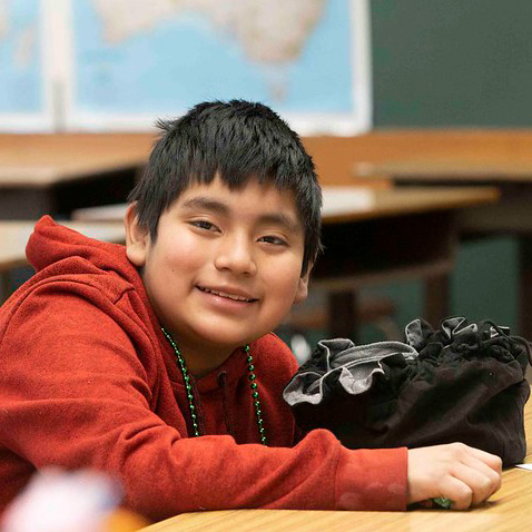A boy in a red sweatshirt sits at a classroom desk, smiling toward the camera.