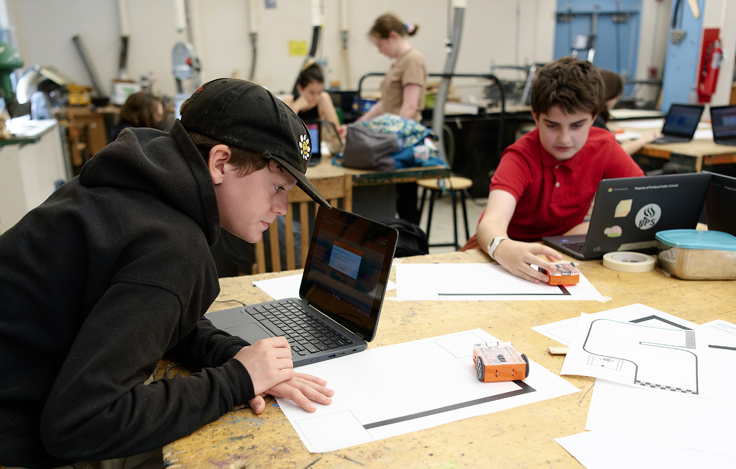 Middle-schoolers program small orange robots with laptops, testing them on paper tracks in a workshop classroom