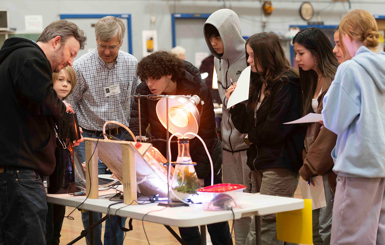 Students gather around a table as mentors demonstrate a science setup with a heat lamp, tubing, and a beaker terrarium